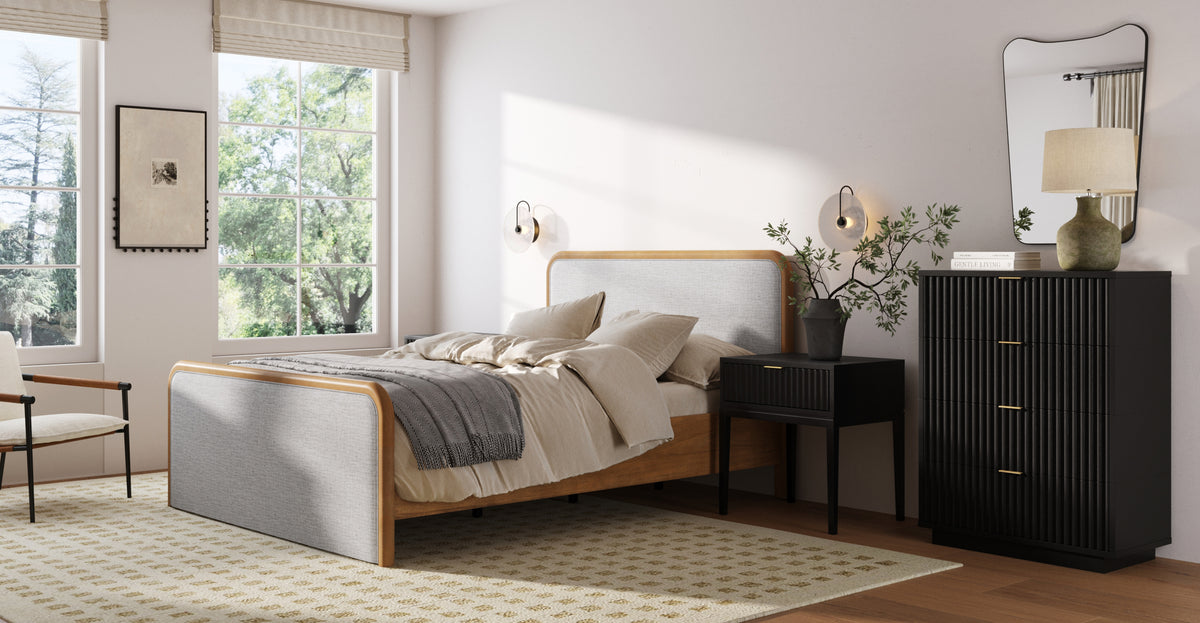 A bedroom featuring a light wood bed frame with grey upholstery, a black fluted nightstand and dresser, a textured area rug, and a mirror leaning against the wall.