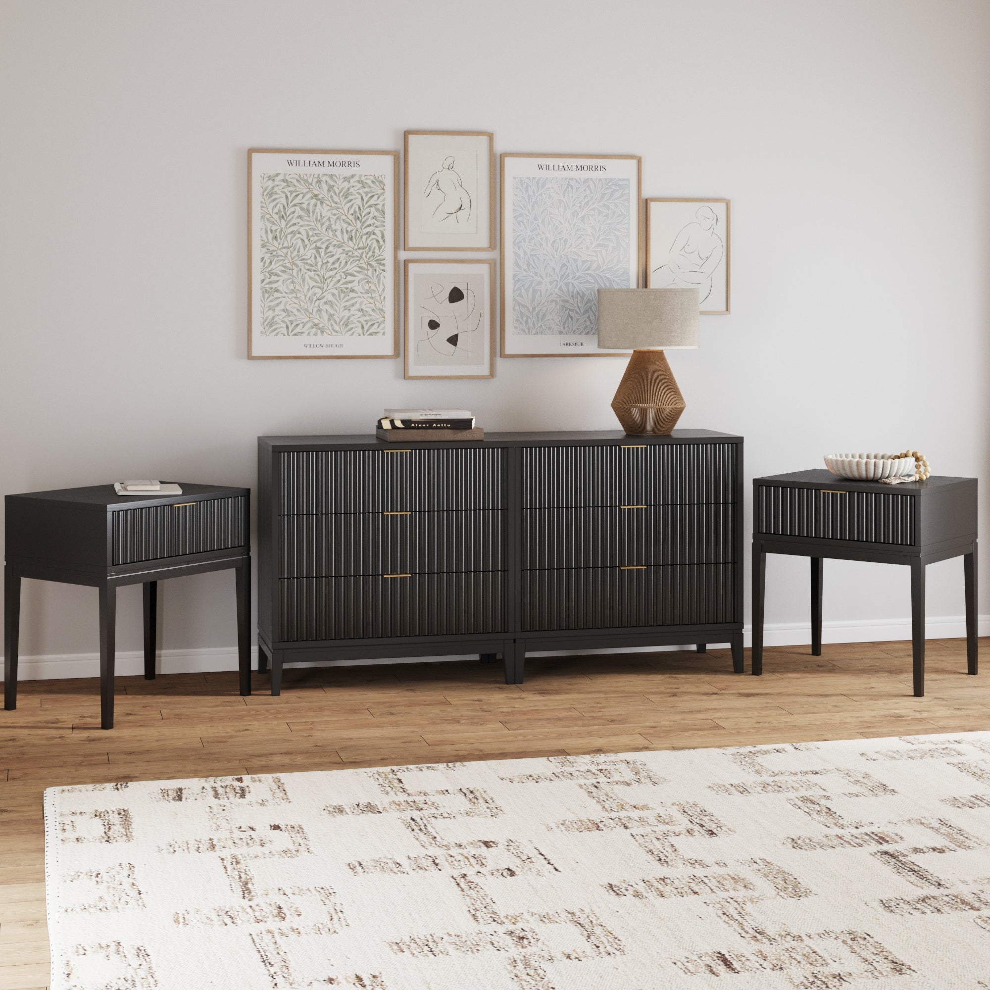 A modern living room with a black sideboard and two matching end tables, all with vertical line patterns. Framed art and a lamp sit on the sideboard. The room has light walls, wood flooring, and a patterned rug.
