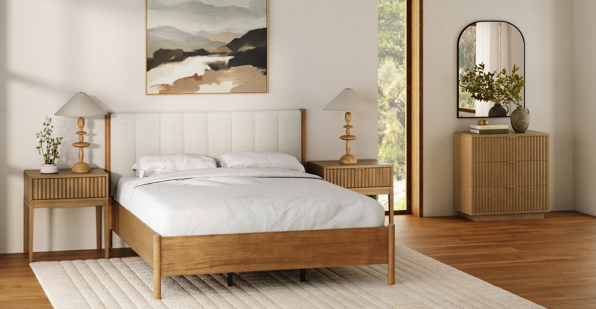 A bedroom featuring a wooden bed frame with a textured white headboard, flanked by two matching wooden nightstands with table lamps, and a wooden dresser with an arched mirror.