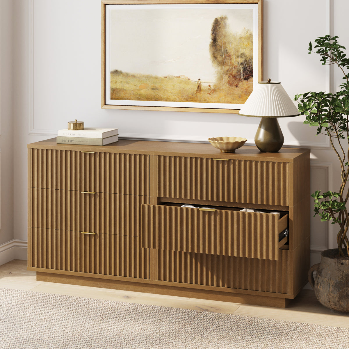 A wooden sideboard with fluted drawer fronts and gold handles, one drawer slightly open, topped with books, a small bowl, and a lamp under a landscape painting.