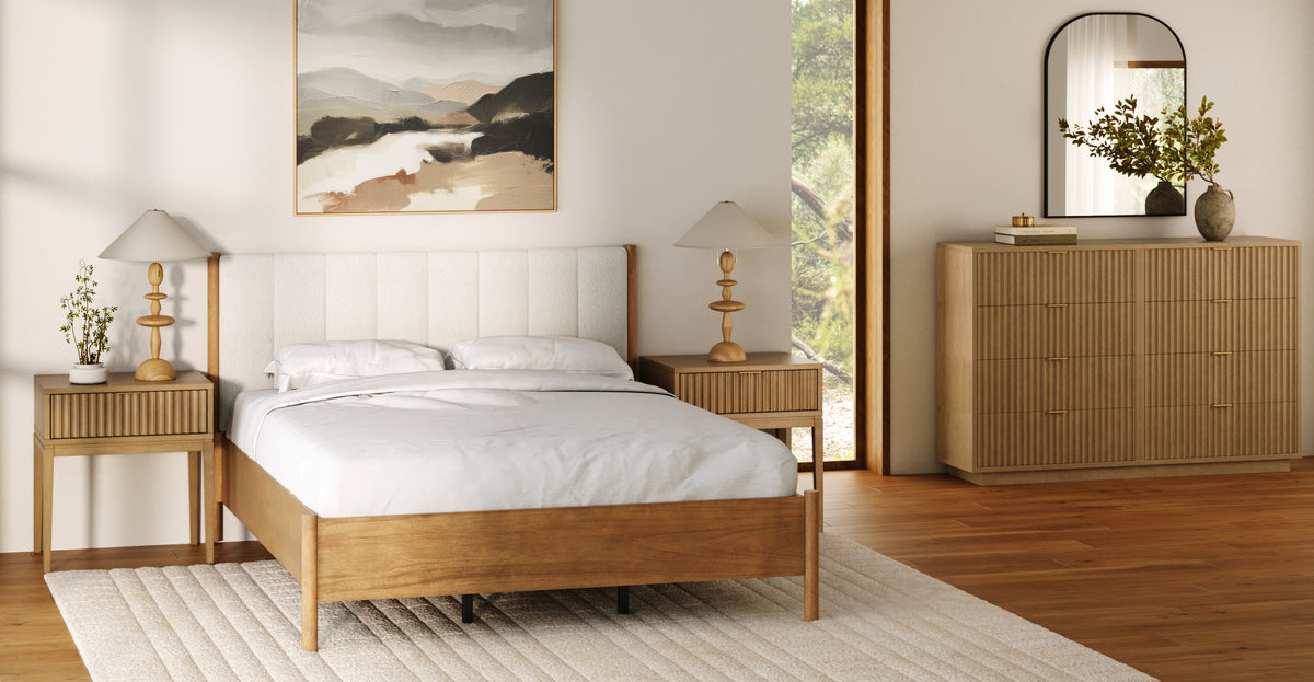 A wooden bed with a white upholstered headboard sits between two matching nightstands, each topped with a wooden table lamp, in a bedroom with a dresser and a wall mirror.