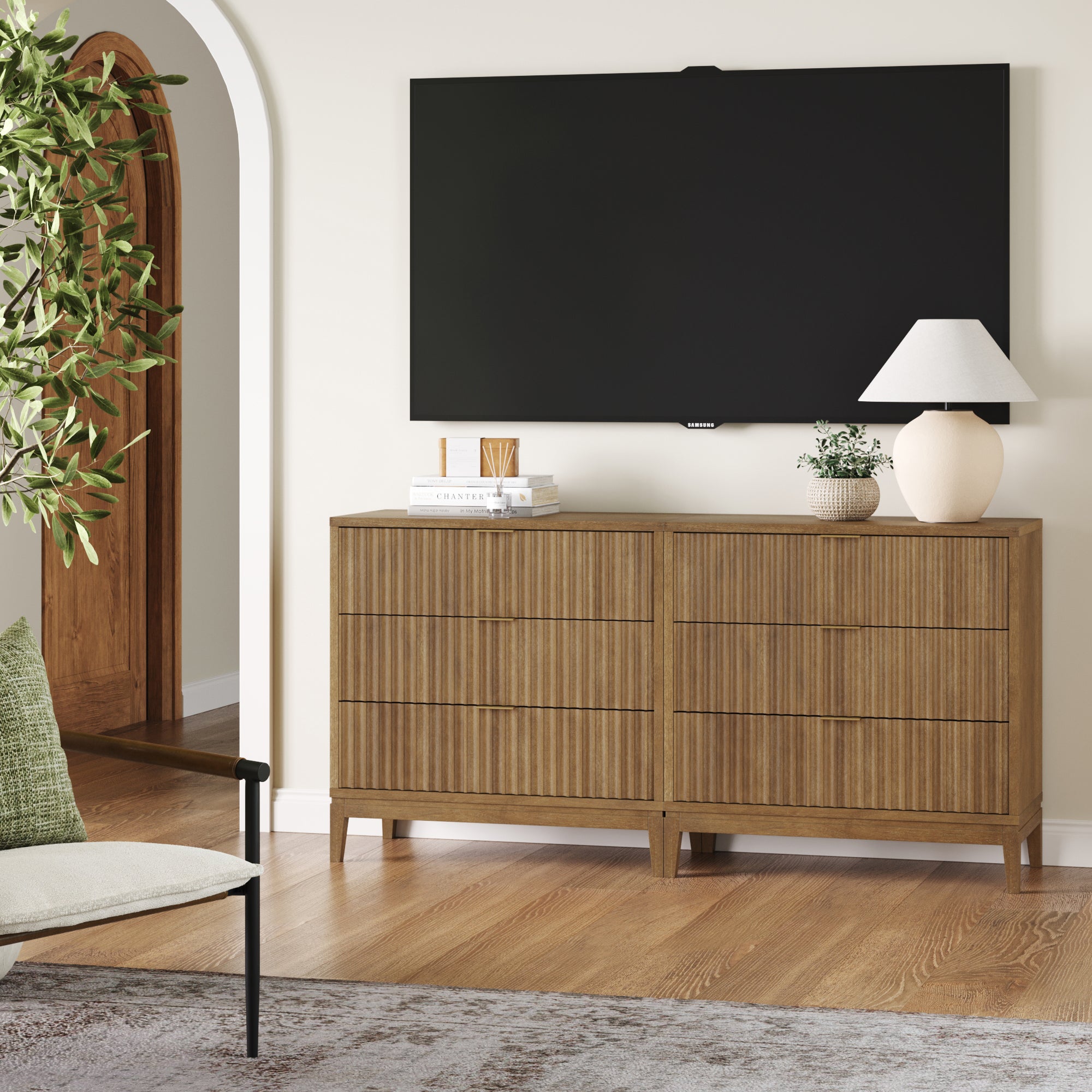 A modern living room with a wooden dresser, a white table lamp, a potted plant, and decor items beneath a wall-mounted flat-screen TV. A green-leafed plant and an archway are visible in the background.