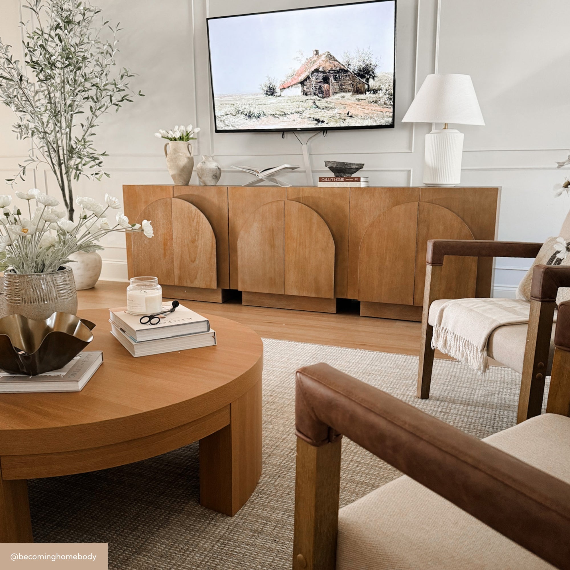 A modern living room with light wood furniture, including a round coffee table and sideboard, beige armchairs, neutral decor, and a wall-mounted TV displaying a cottage painting. Soft lighting and cozy accents complete the space.