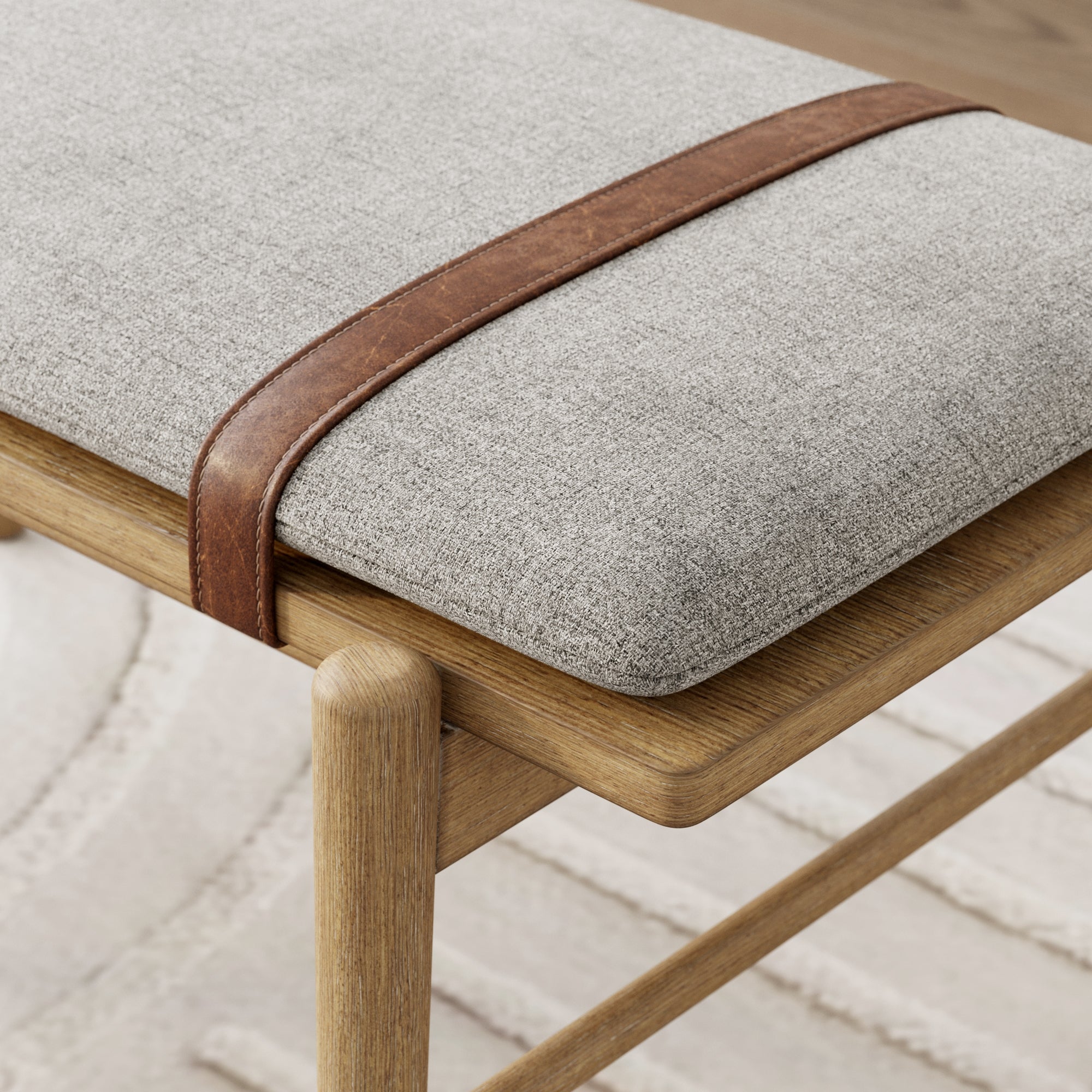 A close-up of a wooden bench with light gray upholstered cushion, featuring a brown leather strap detail across the corner, placed on a textured, cream-colored rug.