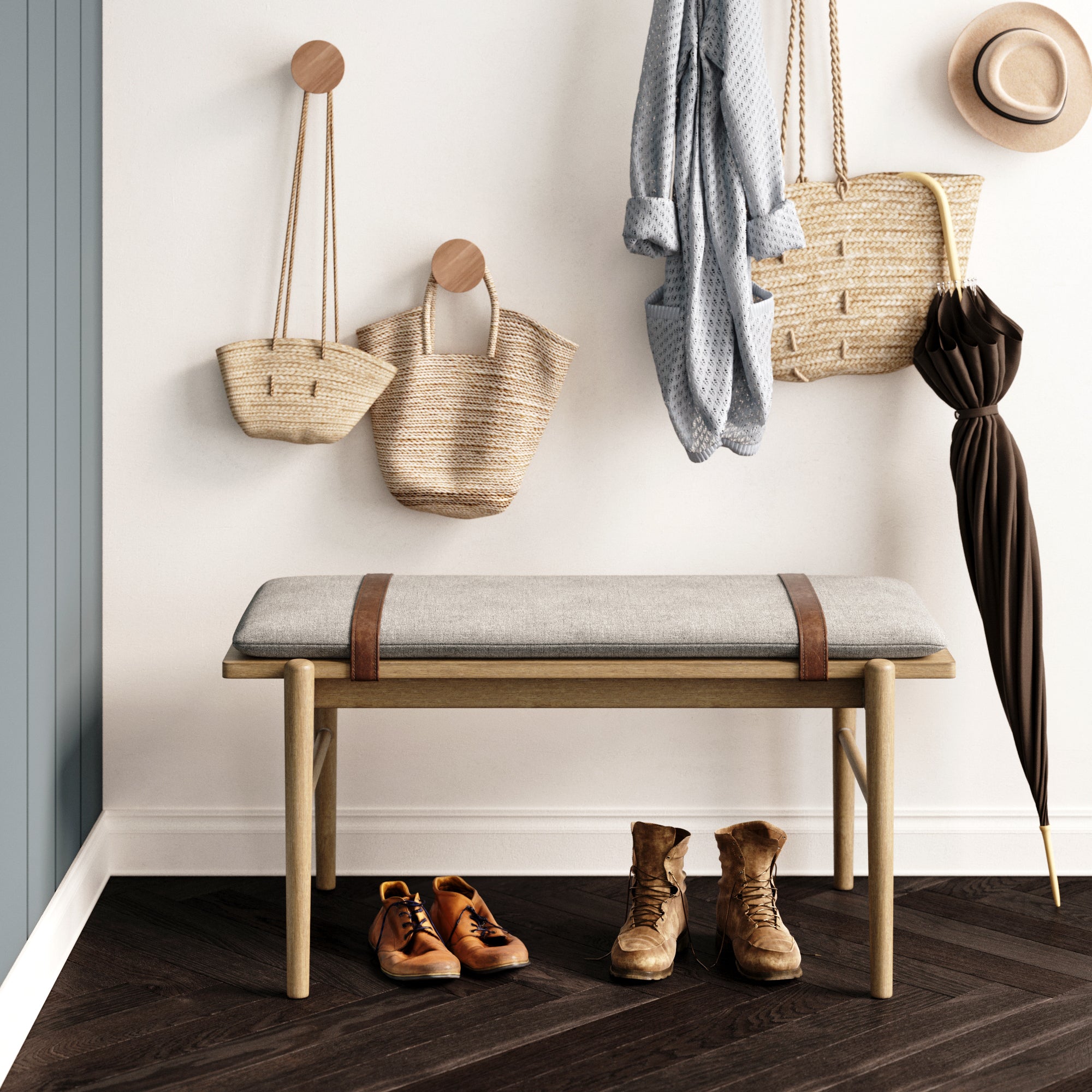 A cozy entryway with a wooden bench, gray cushion, boots, sneakers, woven bags, a hanging coat, an umbrella, and a straw hat on wall hooks, all set against a white wall and dark herringbone floor.