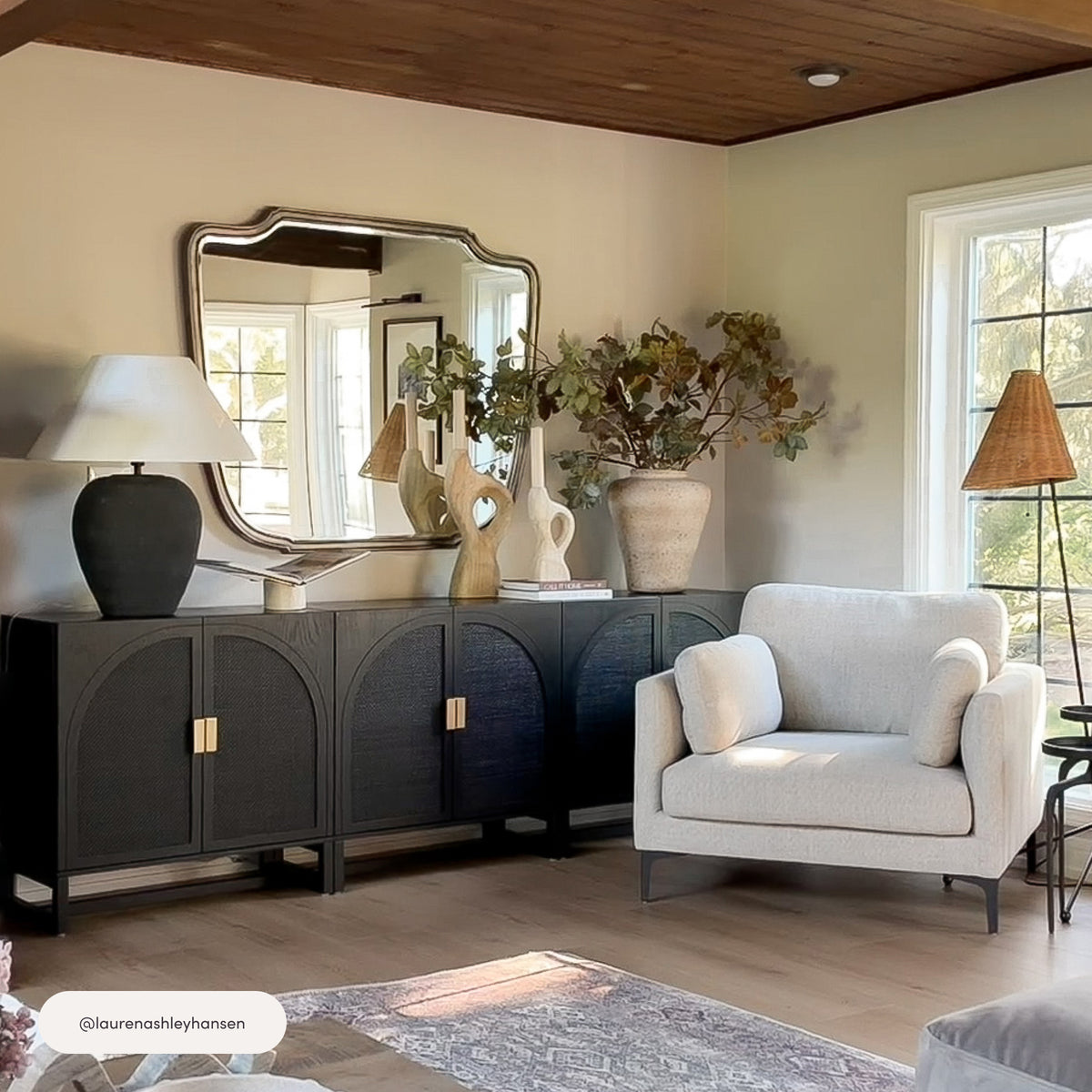 A long, black Nathan James Arched Wooden 2-Door Storage Cabinet Black Rattan with four arched doors and gold handles sits against a wall topped by a large, wavy-edged mirror.