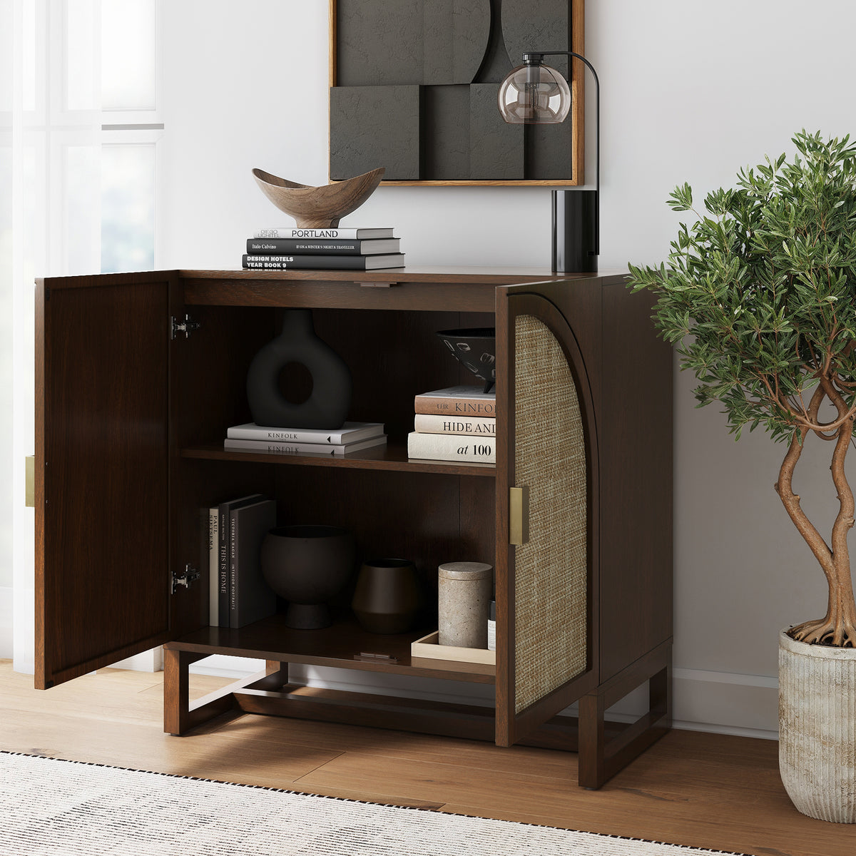 A dark wood cabinet with open doors revealing shelves filled with books and decorative vases, topped with a stack of books and a lamp.