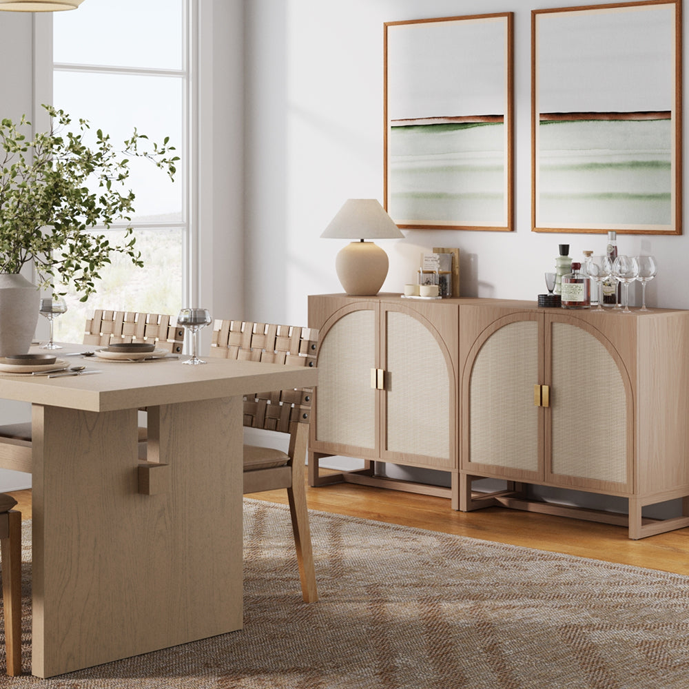A light wood dining table with woven chairs next to a matching arched-door sideboard featuring two abstract framed paintings above it.