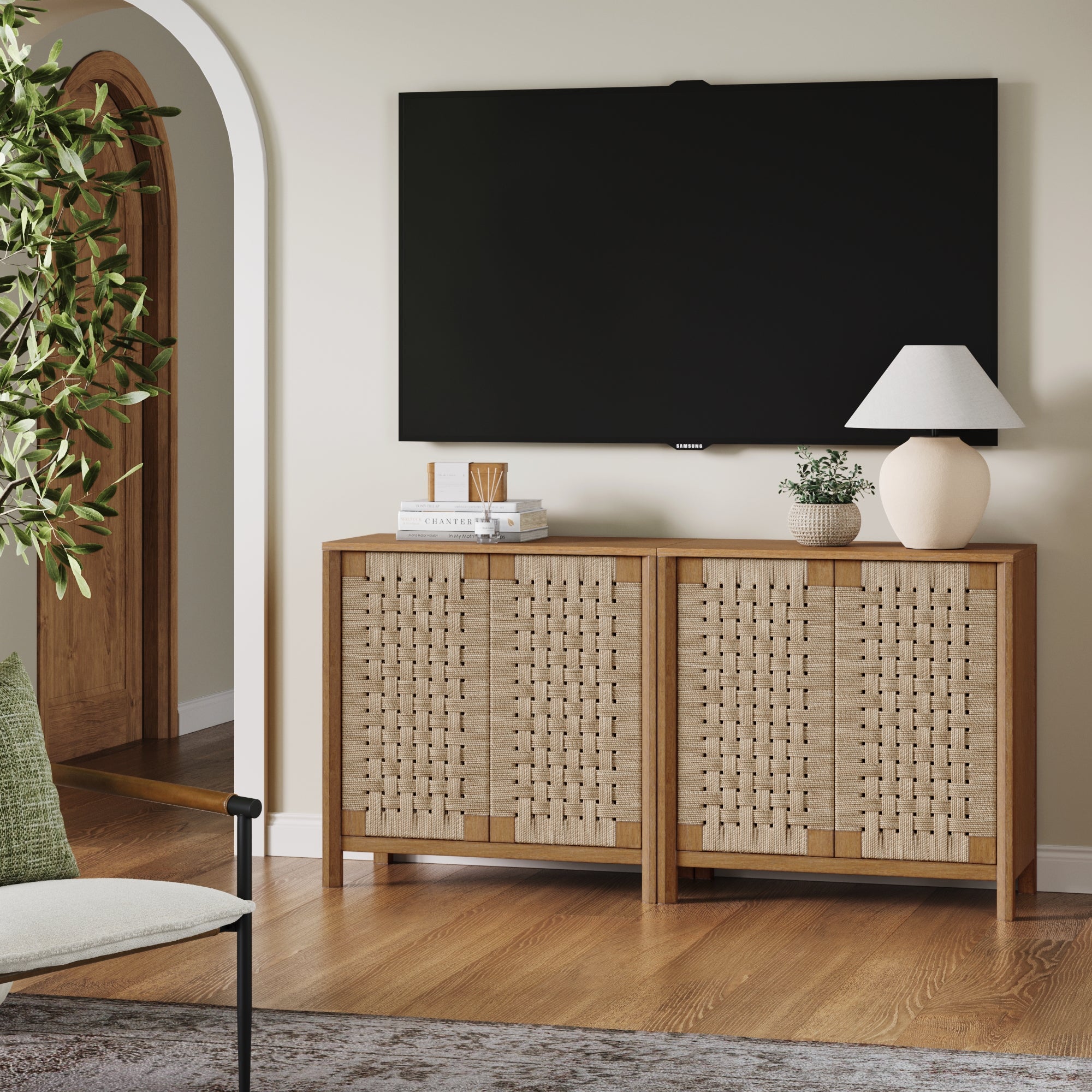 A modern living room with a large flat-screen TV mounted on a light wall above a wooden sideboard with woven doors. A lamp, potted plant, and decor items sit on the sideboard. There is a chair and a green plant nearby.
