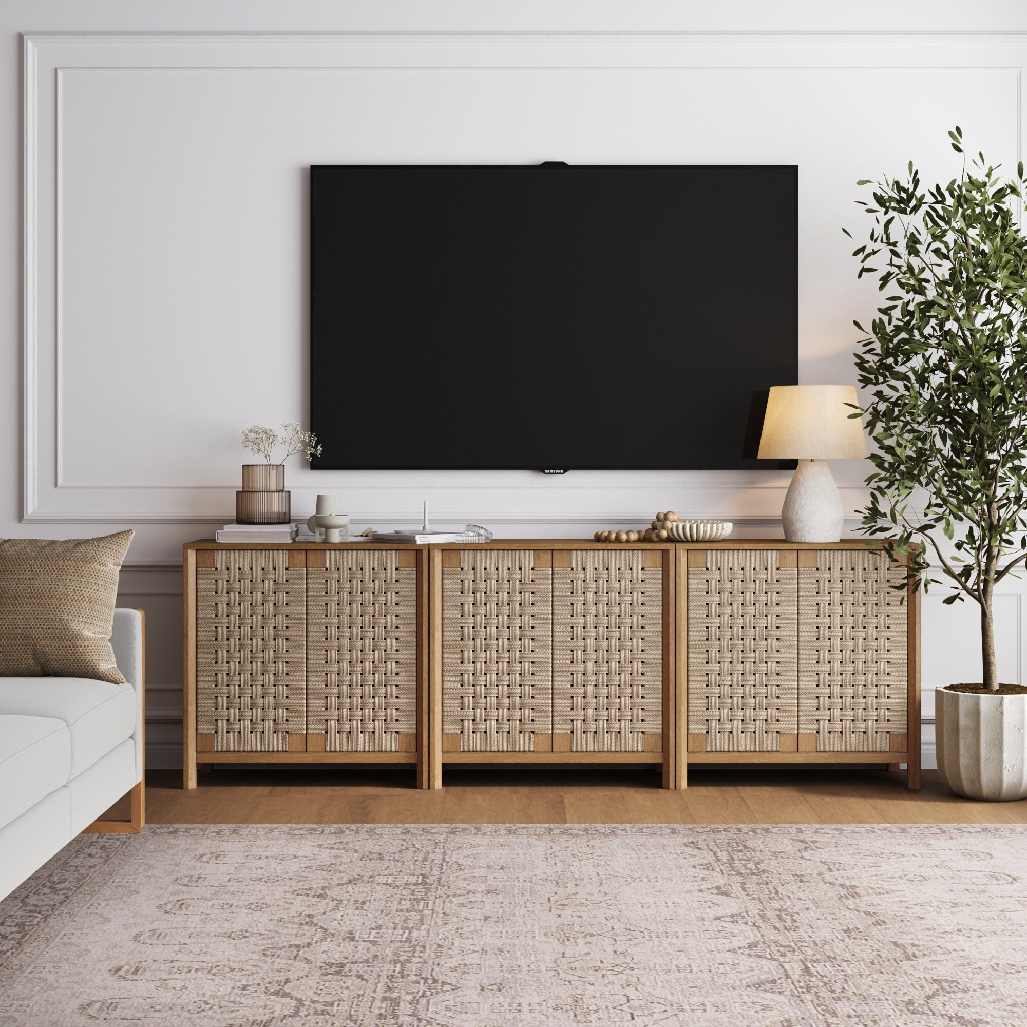 A modern living room with a large flat-screen TV mounted on a white wall, a woven sideboard below it, a table lamp, decor items, a potted tree, and a patterned rug on a wooden floor.