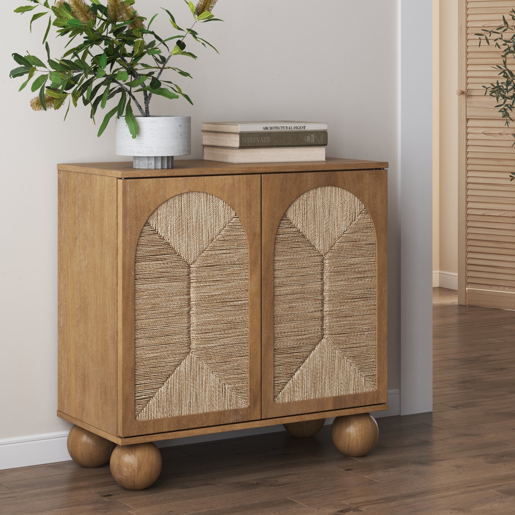 A wooden cabinet with round legs and textured woven front panels stands against a wall. A potted plant and a stack of books sit on top. The floor is wood, and part of a door and hallway are visible in the background.