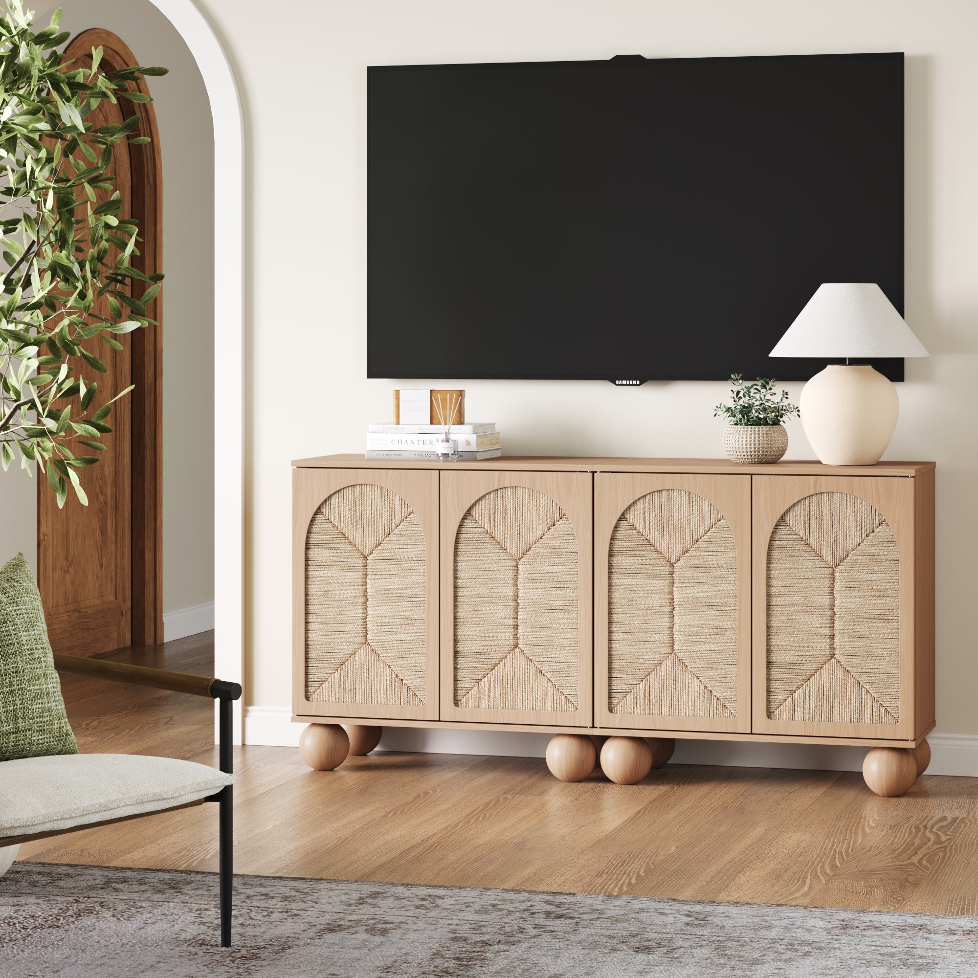 A modern living room with a flat-screen TV mounted on a cream wall above a light wood cabinet with textured doors. Decor includes a lamp, books, a plant, and a chair with a green pillow on a wood floor.