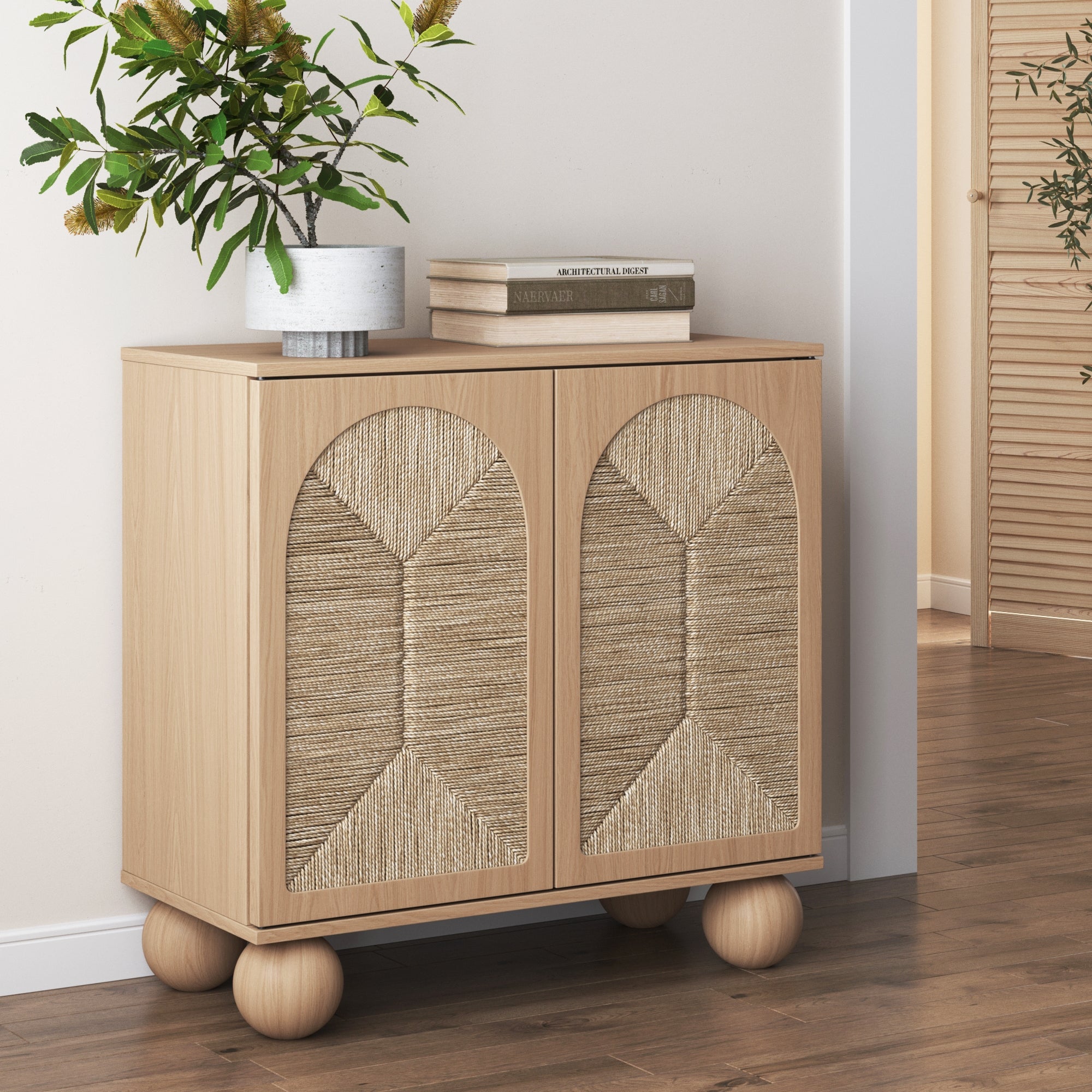 A light wood cabinet with textured, woven doors and round legs stands against a wall. A potted plant and a stack of books decorate the top; a doorway and additional wood panels are visible in the background.
