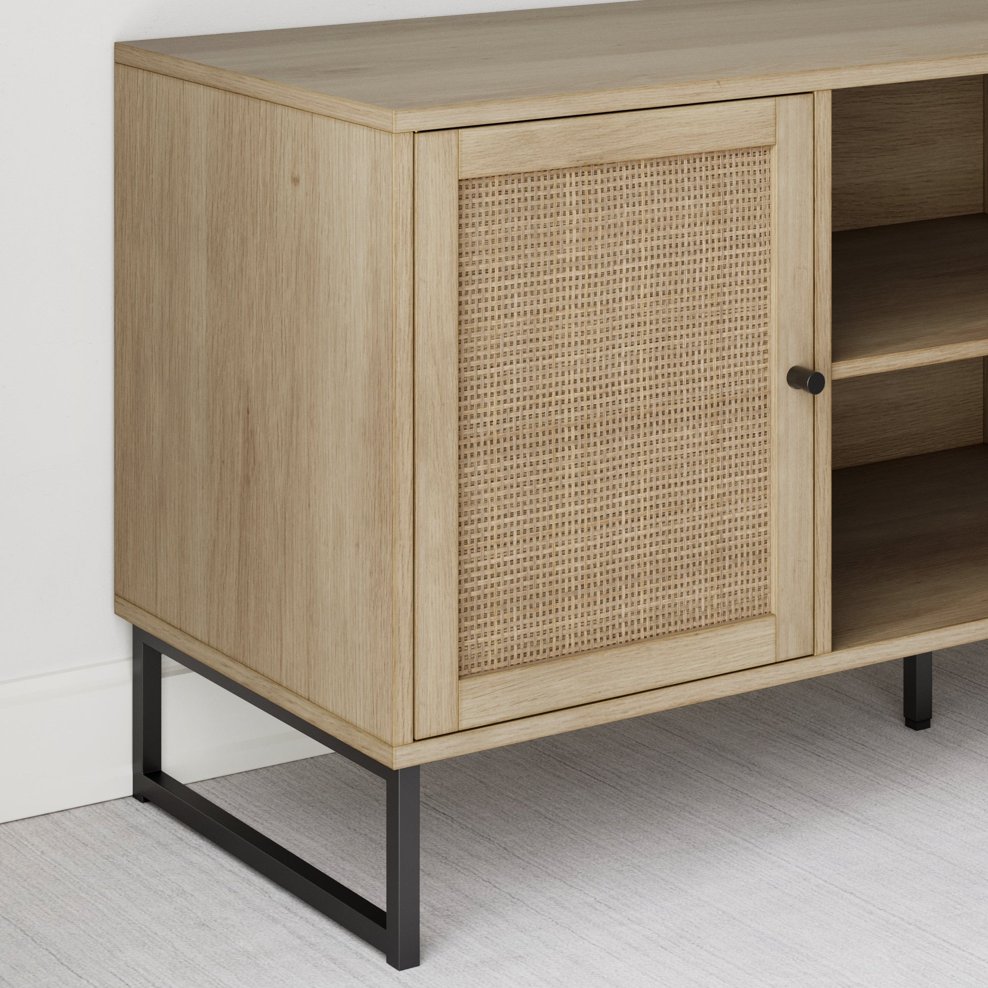 A light wood cabinet with a woven rattan door, open shelving, and black metal legs, placed against a white wall on a light-colored floor.