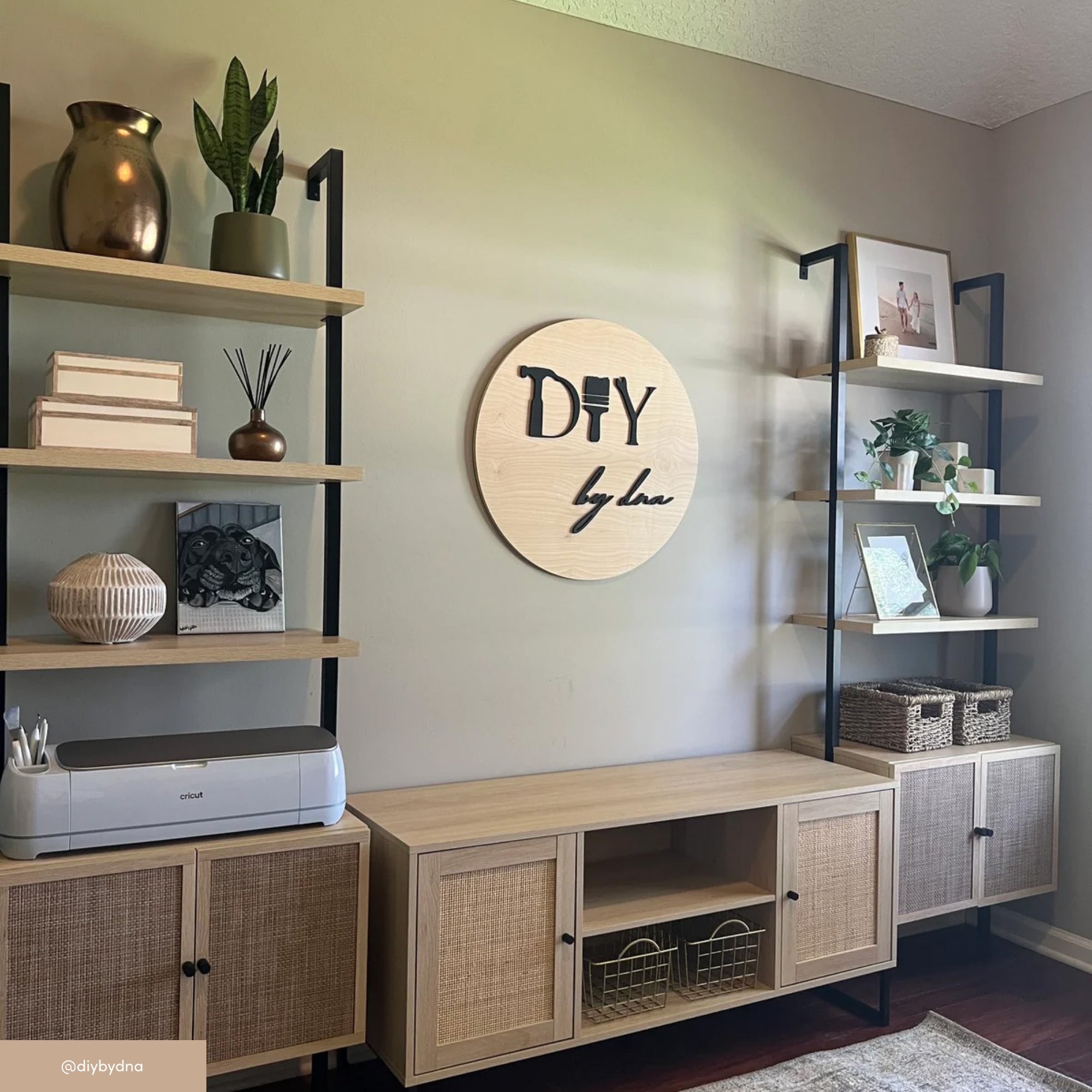 A modern home office with light wood and black shelving units, woven baskets, potted plants, books, decor, and a crafting machine. A round wooden sign on the wall reads DIY by Luv.