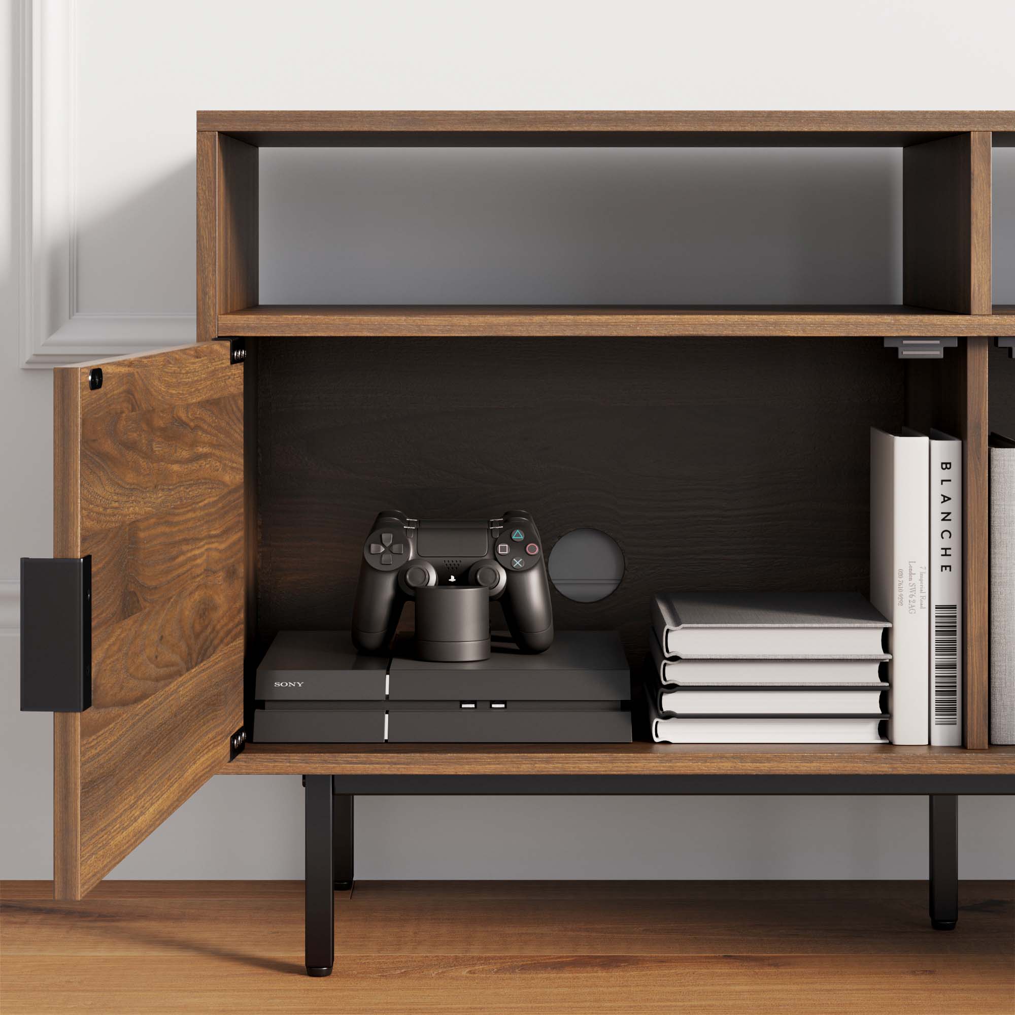 A wooden media console with an open cabinet door reveals a PlayStation console, two controllers, stacked books, and a few vertical books inside. The cabinet has black metal legs and sits on a wood floor.