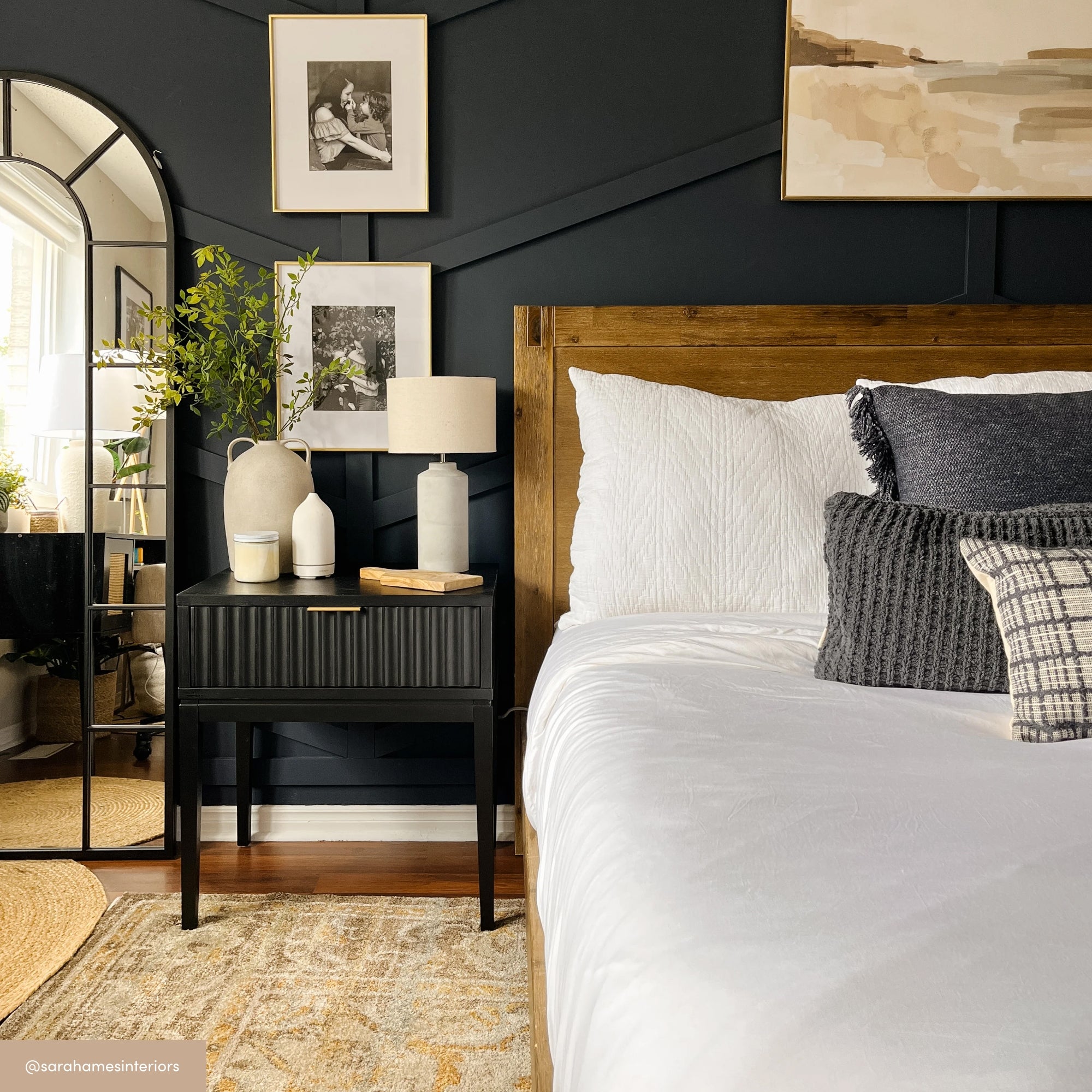 A modern bedroom features a wooden bed with white and gray bedding, a black nightstand with vases and a lamp, a large arched mirror, wall art, and dark accent walls. A rug and indoor plant complete the cozy decor.