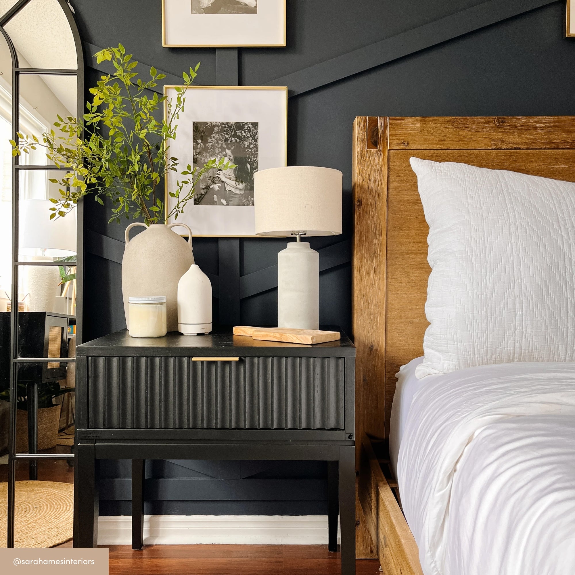 A modern bedroom features a wooden bed with white bedding, a black nightstand with a drawer, a beige lamp, decorative vases, greenery, candles, wall art, and a tall arched mirror against a dark accent wall.
