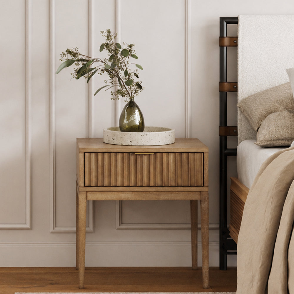 Wooden nightstand with a fluted drawer front, topped with a round tray holding a small glass vase with leafy branches, positioned next to a bed.