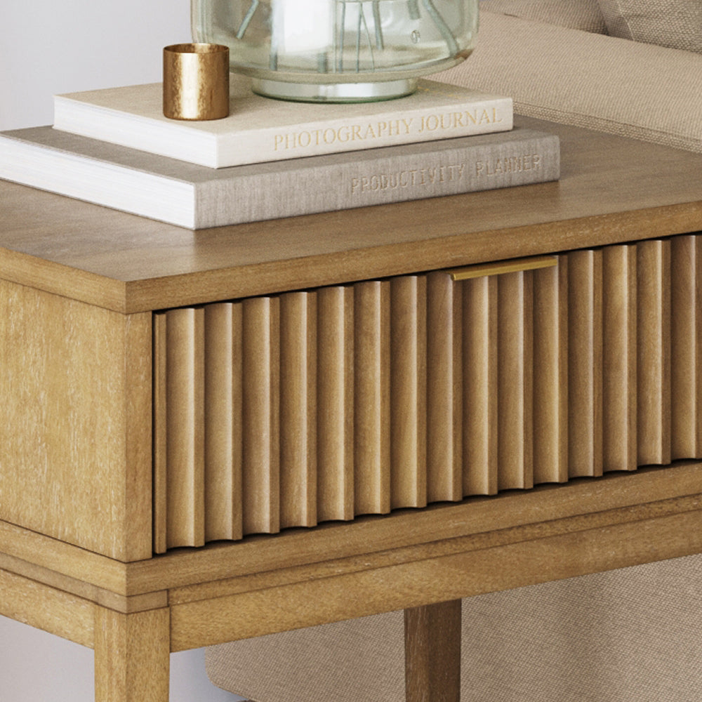 Light wood nightstand with a fluted drawer front, topped with two books, a gold candle, and a glass vase.