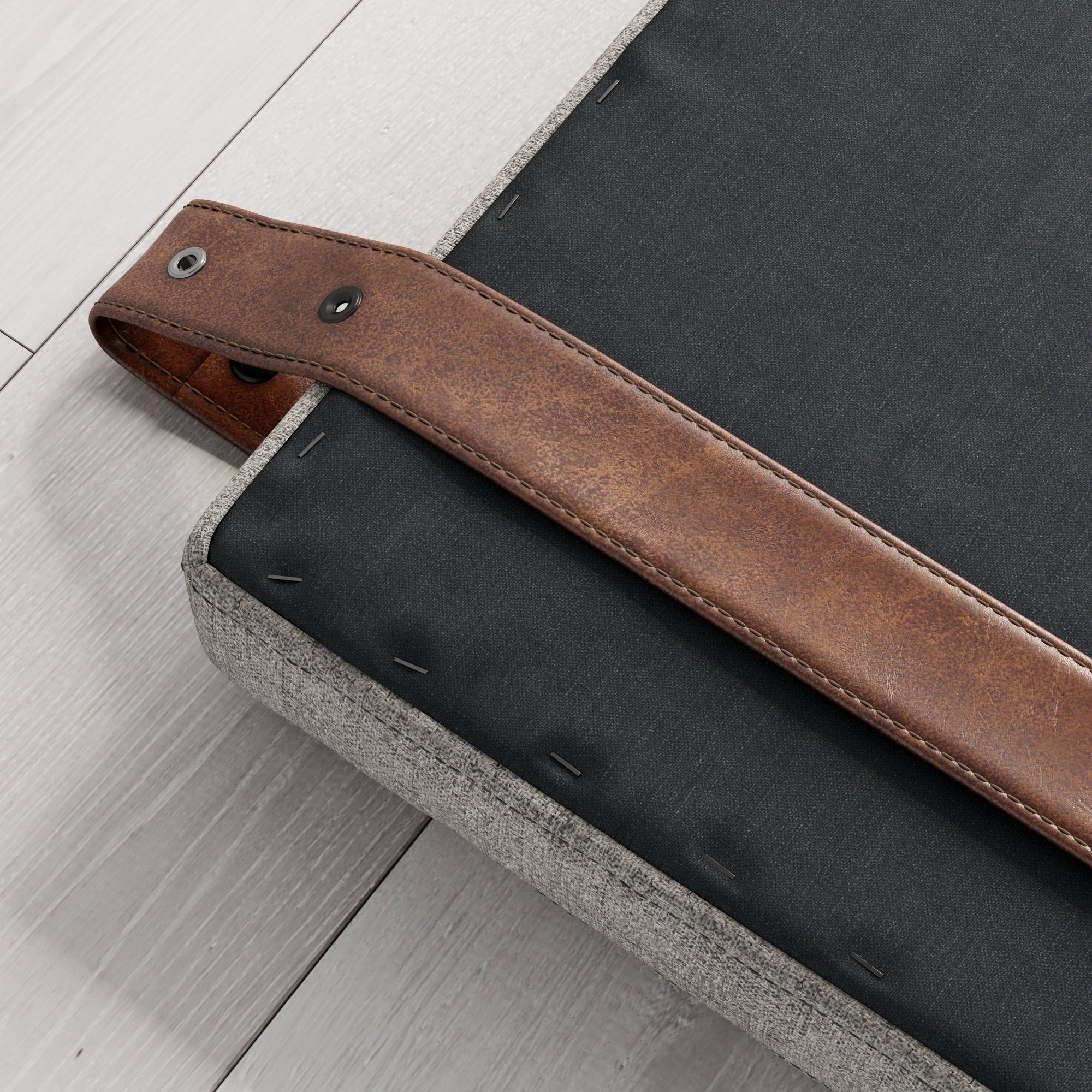 A close-up of a gray upholstered furniture piece with a black base and a brown leather strap attached, secured by metal staples and a rivet, resting on light-colored wooden flooring.