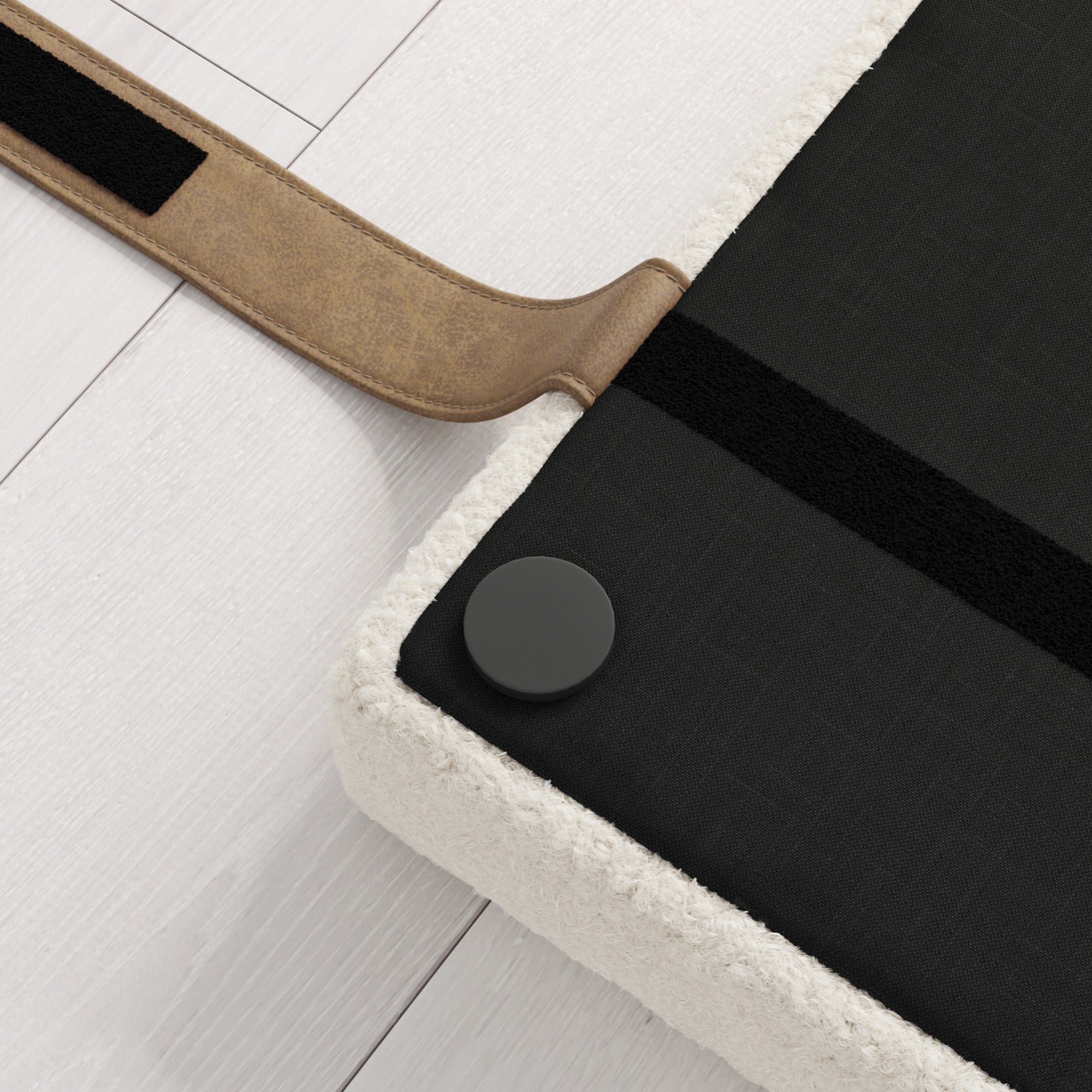 Close-up of a black cushioned surface with a beige fabric edge, a brown leather strap, and a round black button, placed on light-colored wooden flooring.