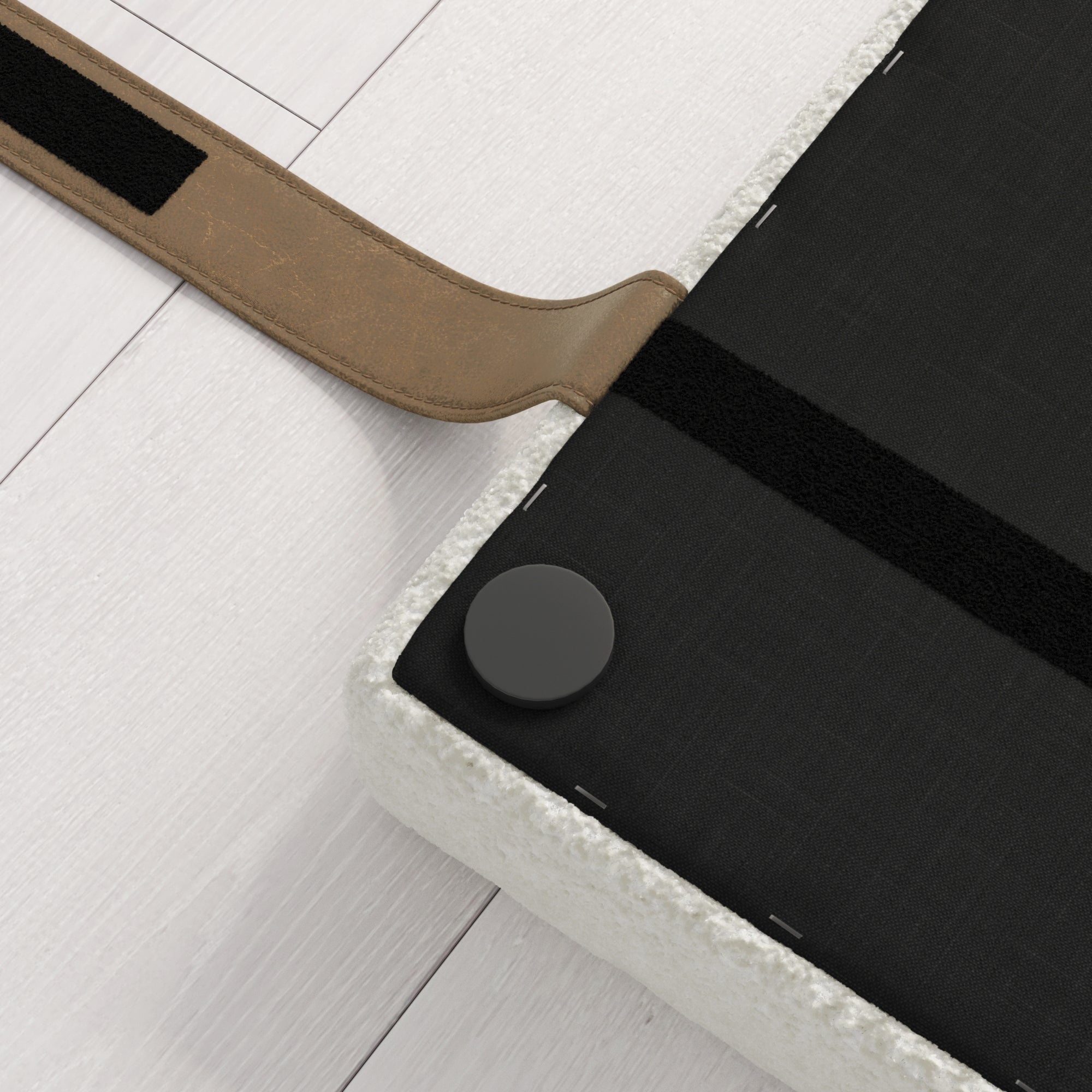 A close-up view of a mattress corner with a black bottom cover, a round black button, and a brown strap attached, placed on a white wooden floor.