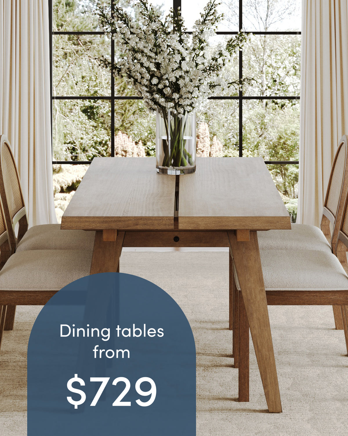 A wooden dining table and two chairs in front of a window, with a glass vase of white flowers on the table and a graphic overlay that reads, Dining tables from $729.