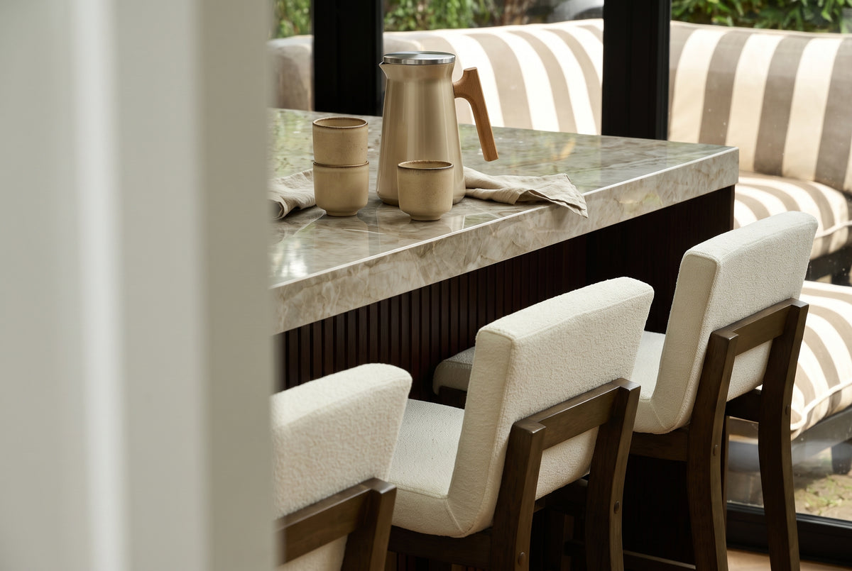 A pitcher and three small cups sit on a stone kitchen island with three wooden bar stools in the foreground.