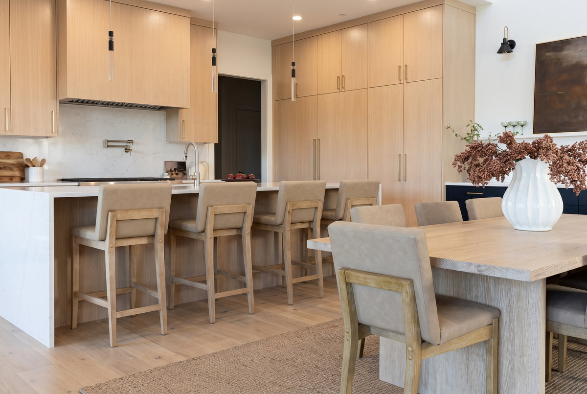 A modern kitchen featuring light wood cabinetry, a waterfall island with four bar stools, and a dining area with a wooden table and matching upholstered chairs.