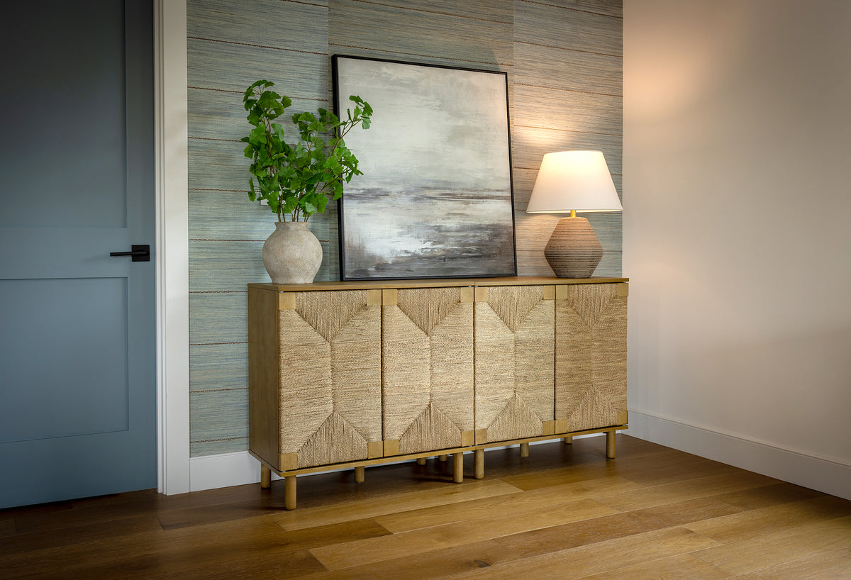A wooden sideboard with four woven doors stands against a textured wall, topped with a potted plant, an abstract framed painting, and a table lamp.