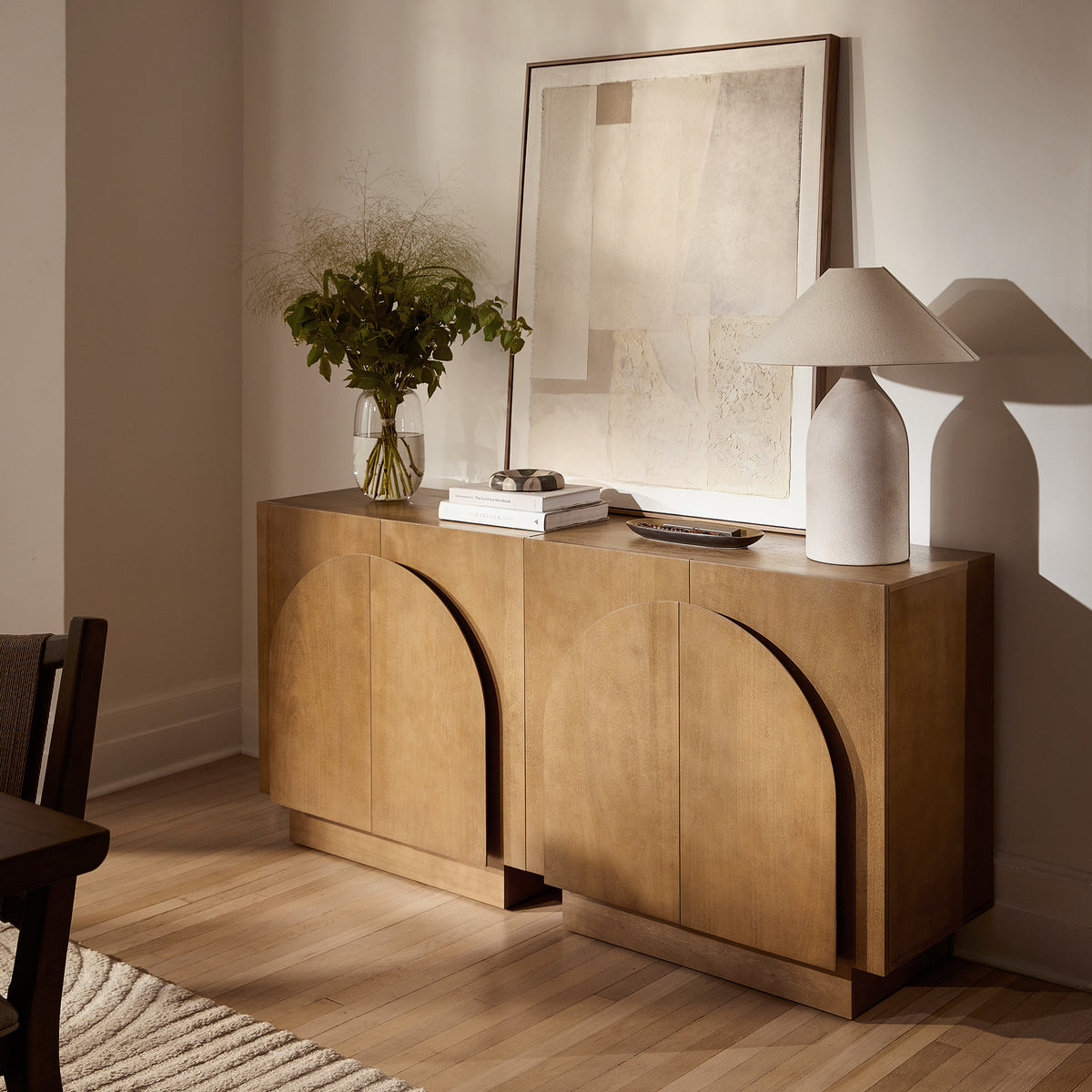 Nathan James Wood Arched TV Console Stand for TVs up to 100″ Brown featuring two arched cabinet doors, styled with a vase of flowers, a framed abstract print, and a table lamp.