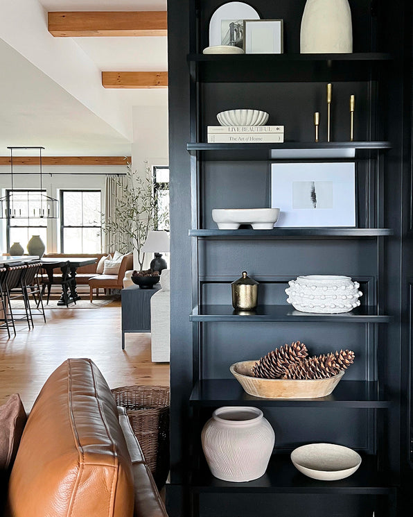 A tall, black bookshelf decorated with various ceramic bowls, vases, pinecones, and books, situated in a living room with a view of an open-concept kitchen and dining area.