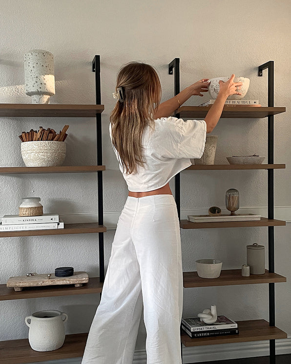 A person in a white top and pants stands before a tall shelving unit, placing a decorative bowl on an upper shelf.