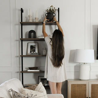 A person stands on their tiptoes to place a dark vase on the top shelf of a metal and wood bookcase.