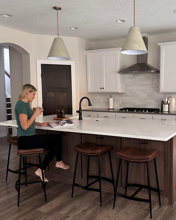 A woman sits at a kitchen island holding a mug and reading a magazine.