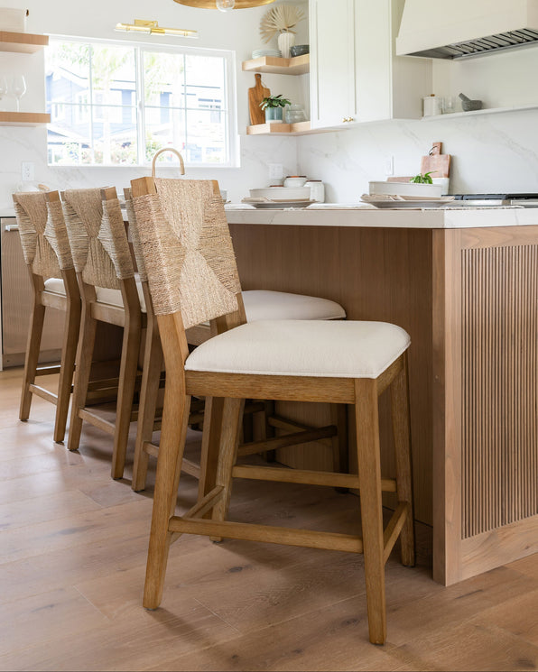 Three wooden counter stools with woven backs and off-white upholstered seats lined up at a kitchen island.