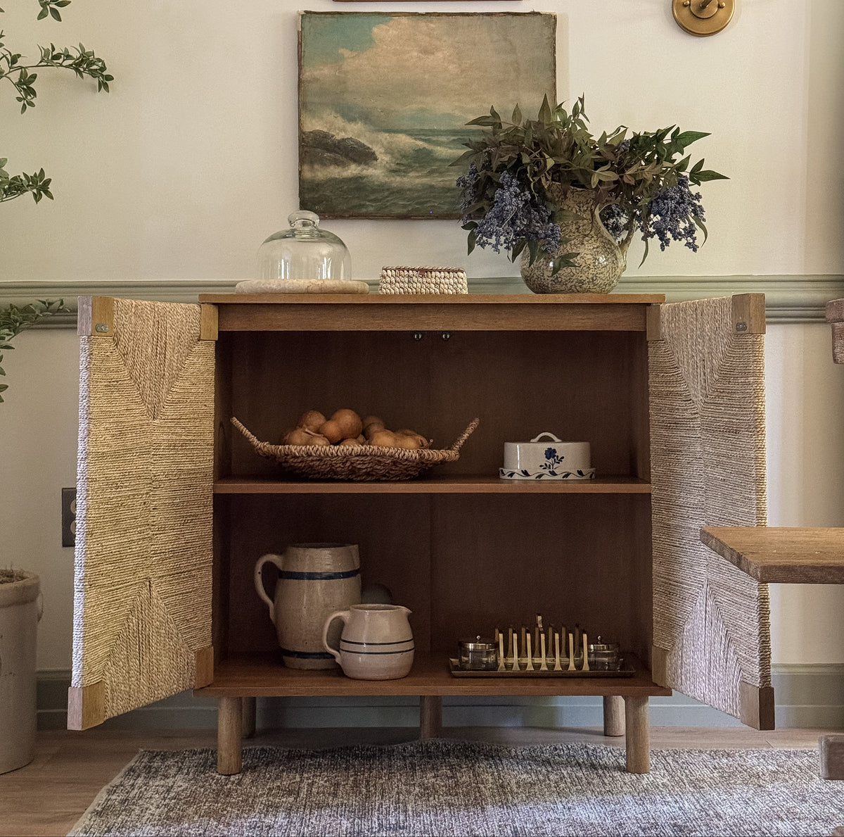 A wooden cabinet with woven doors stands open, revealing shelves holding a basket of onions, a ceramic butter dish, two stoneware pitchers, and a small metal candle holder.