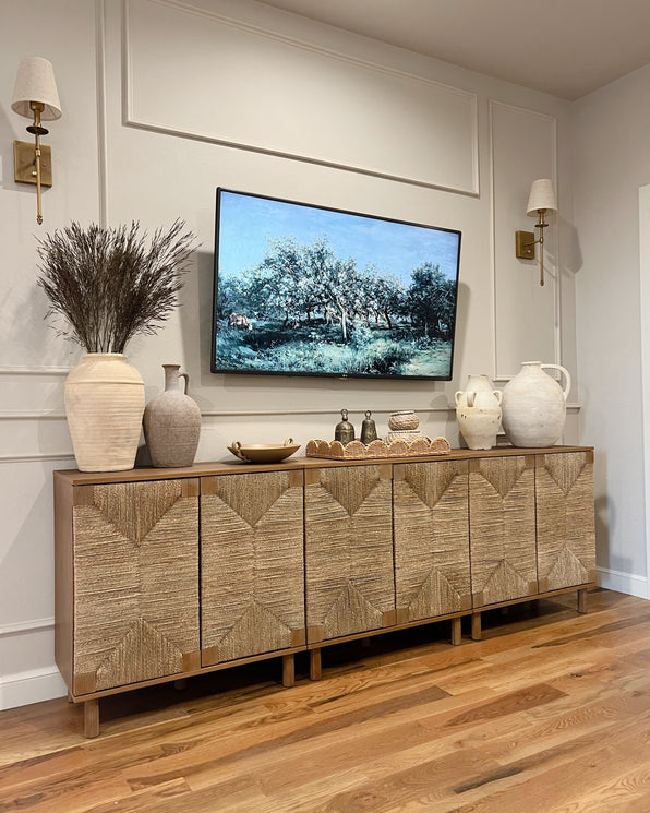 A wooden sideboard with woven textured doors sits below a wall-mounted TV displaying a landscape painting, flanked by two wall sconces and decorated with various ceramic vases and decorative objects.