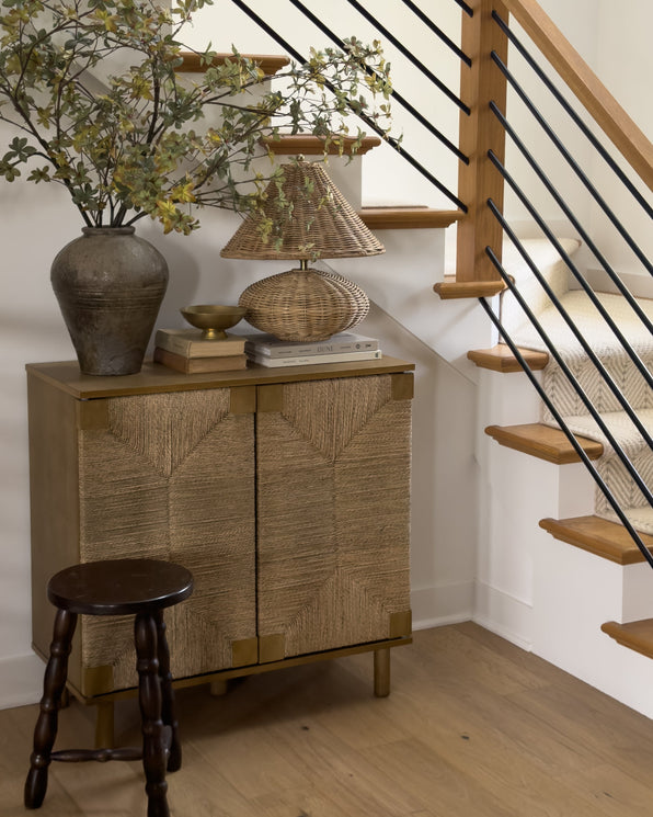 A wooden cabinet with woven doors, topped with a vase of branches, a lamp, and stacked books, next to a small wooden stool at the base of a staircase.