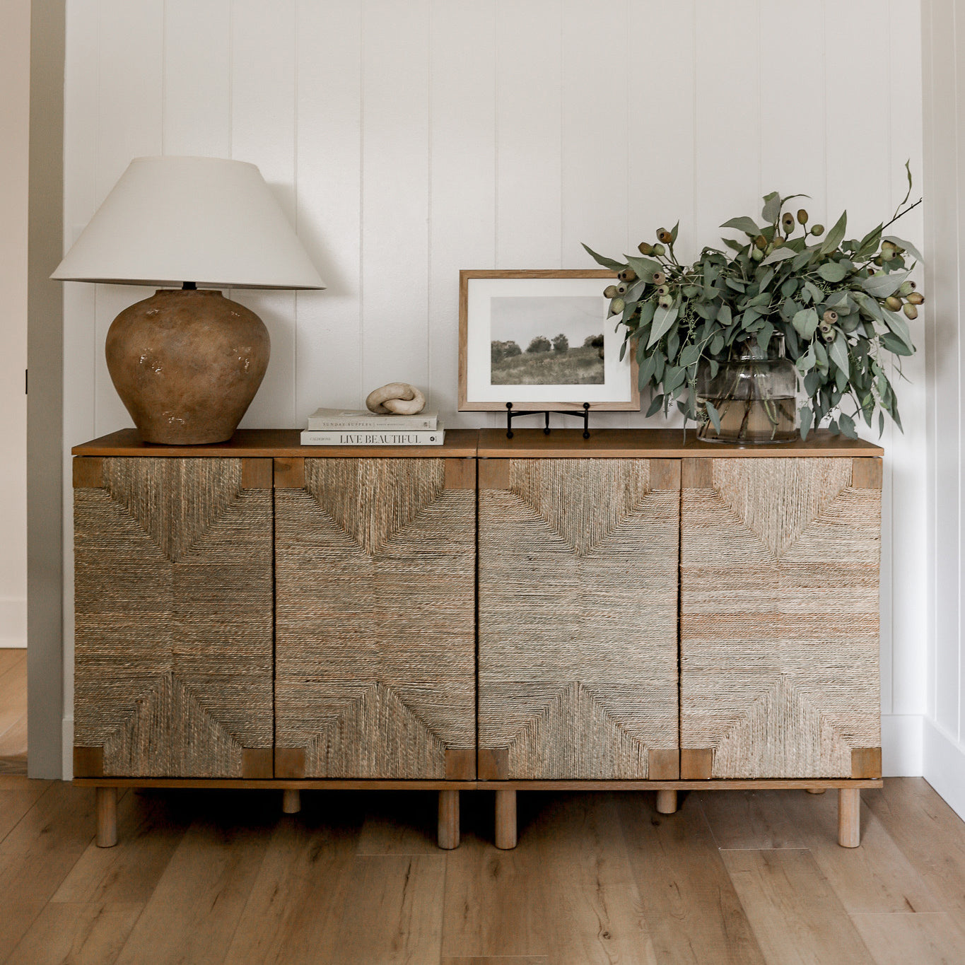 A wooden sideboard with textured doors stands against a white wall. On top are a large ceramic lamp, stacked books, a framed photo, and a vase with leafy green branches. The floor is light wood.
