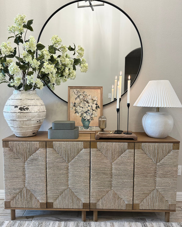 A wooden sideboard with woven textured doors topped with a large vase of flowers, a floral print, three lit taper candles, and a pleated lamp, with a large circular mirror on the wall behind.