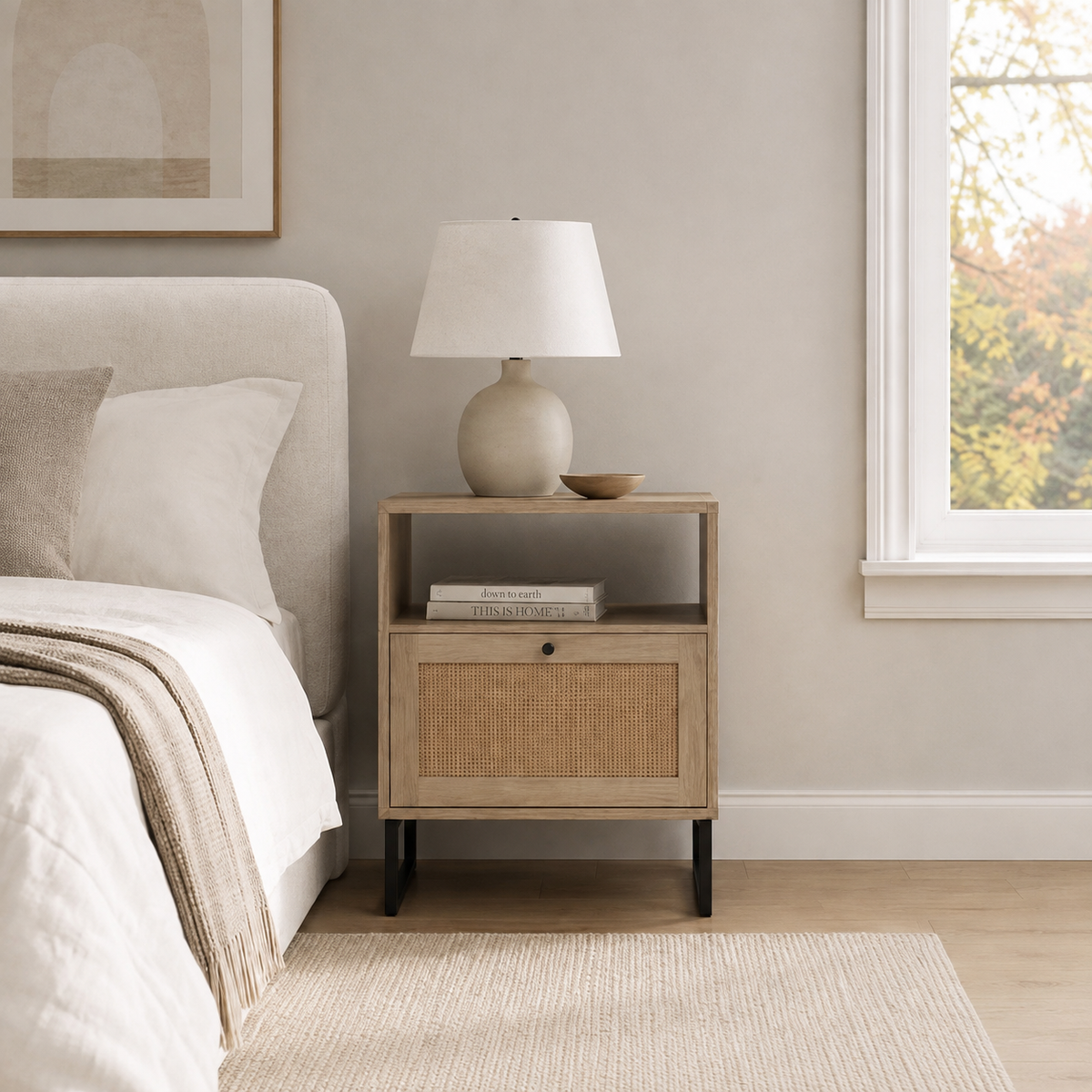 A wooden nightstand with an open shelf and a cane-front drawer, topped with a table lamp and a small bowl, sitting next to a bed.