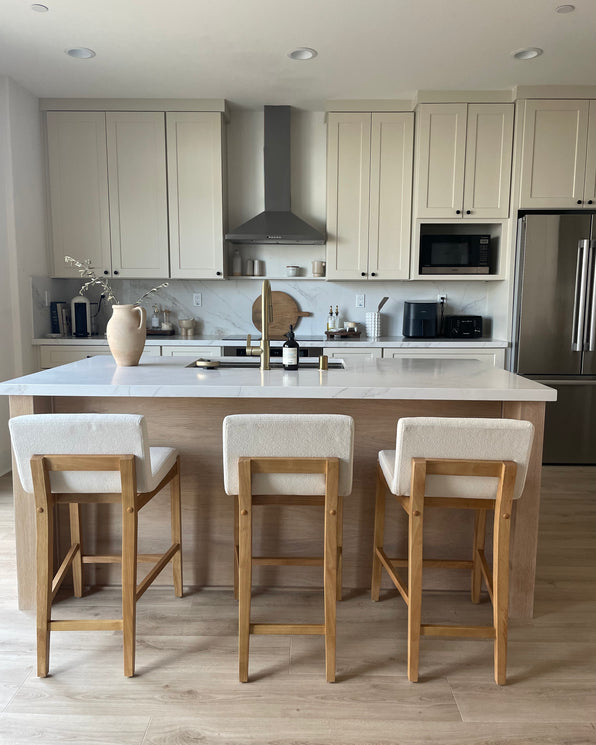 A modern kitchen with a large island featuring three upholstered wooden bar stools and a sink, set against cream-colored cabinets and a stainless steel range hood.