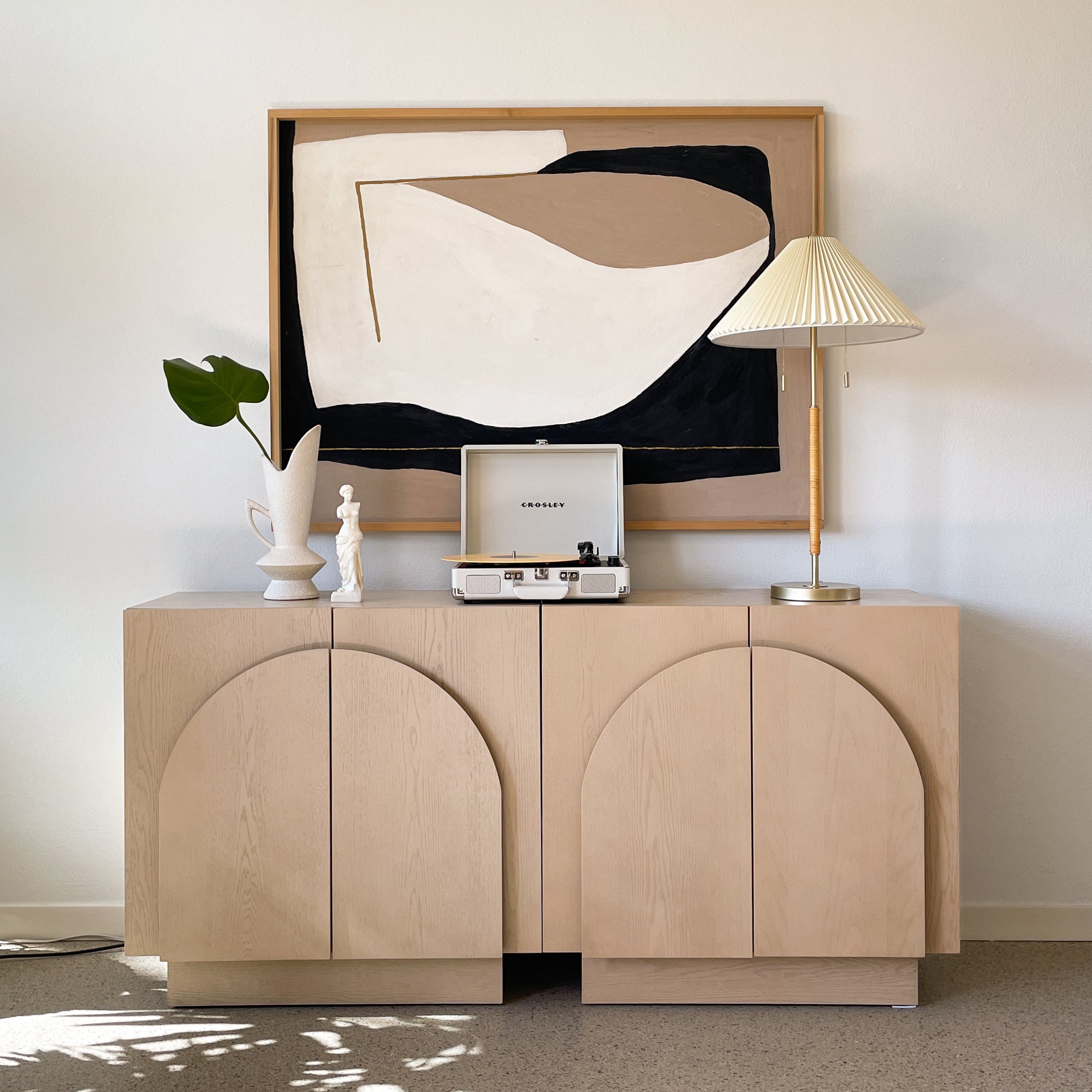 A modern sideboard with arch-shaped doors holds a record player, ceramic vase with a green leaf, a white figurine, and a gold lamp with a pleated shade. An abstract framed artwork hangs on the wall behind.