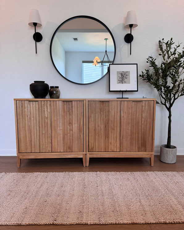 Two side-by-side wooden cabinets topped with two vases, a framed botanical print, and a large round mirror between two wall sconces, with a faux potted tree to the right.
