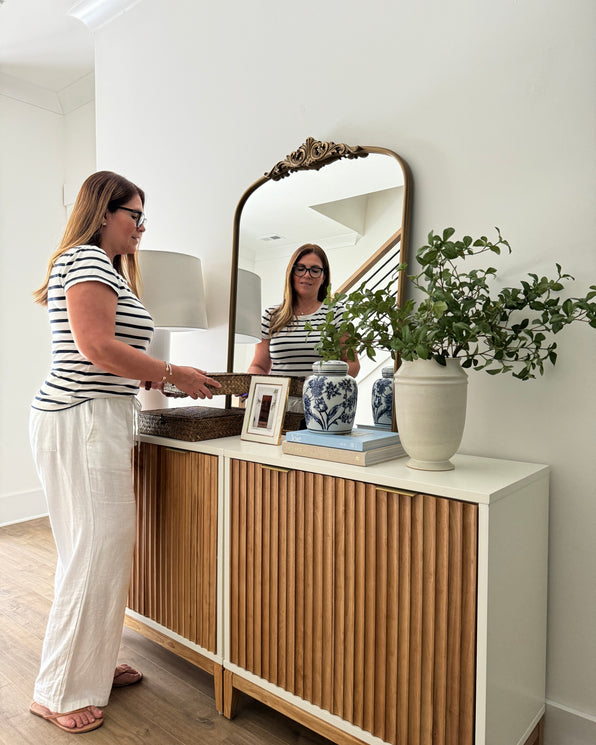 A woman in a striped shirt and white pants places a woven tray on a wooden sideboard topped with a vase, books, and an arched mirror.