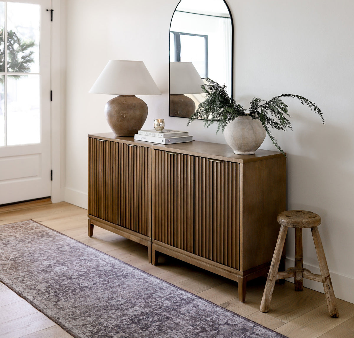 A wooden console table with slatted doors sits against a white wall under an arched mirror, topped with a lamp, books, and a vase with greenery, with a small wooden stool to the side.