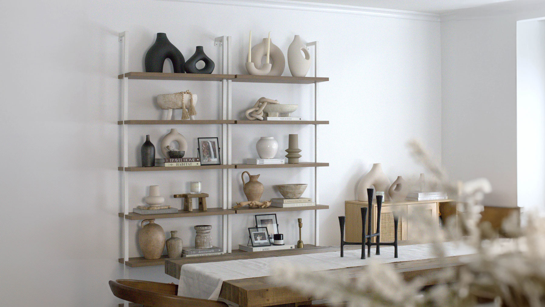 Minimalist dining room with a wooden table, chairs, and a white table runner. In the background, wall-mounted shelves display neutral-toned ceramics, vases, books, and decorative objects. The space is bright and modern.