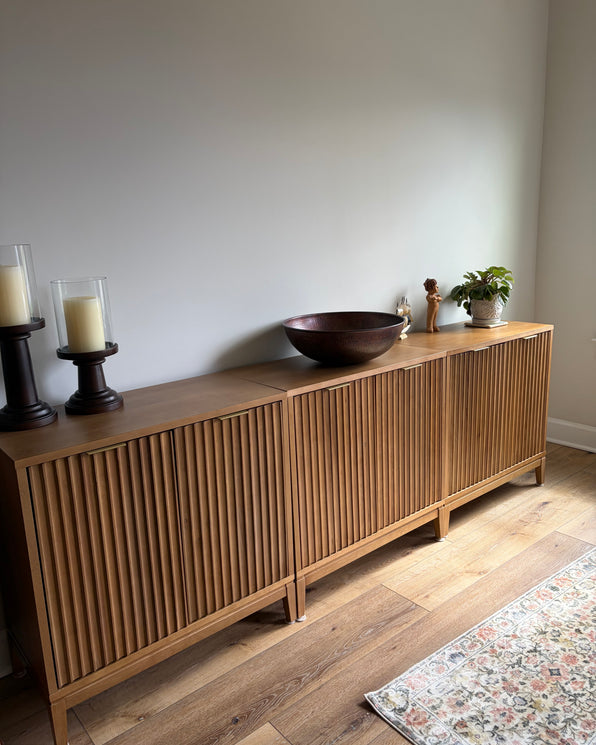 A long wooden sideboard with fluted doors, topped with two candle holders, a large copper-toned bowl, a small figurine, and a potted plant.